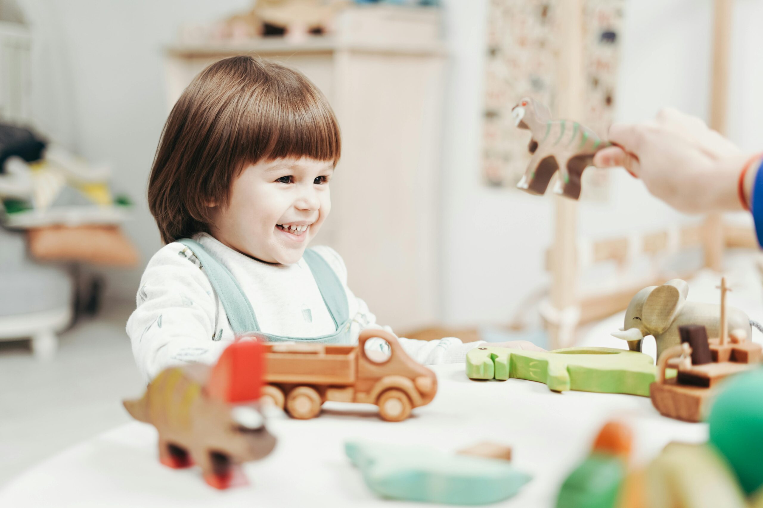 young three year old boy smiling while sitting at a table while he plays with a wooden truck on wheels to meet his play milestones in early childhood