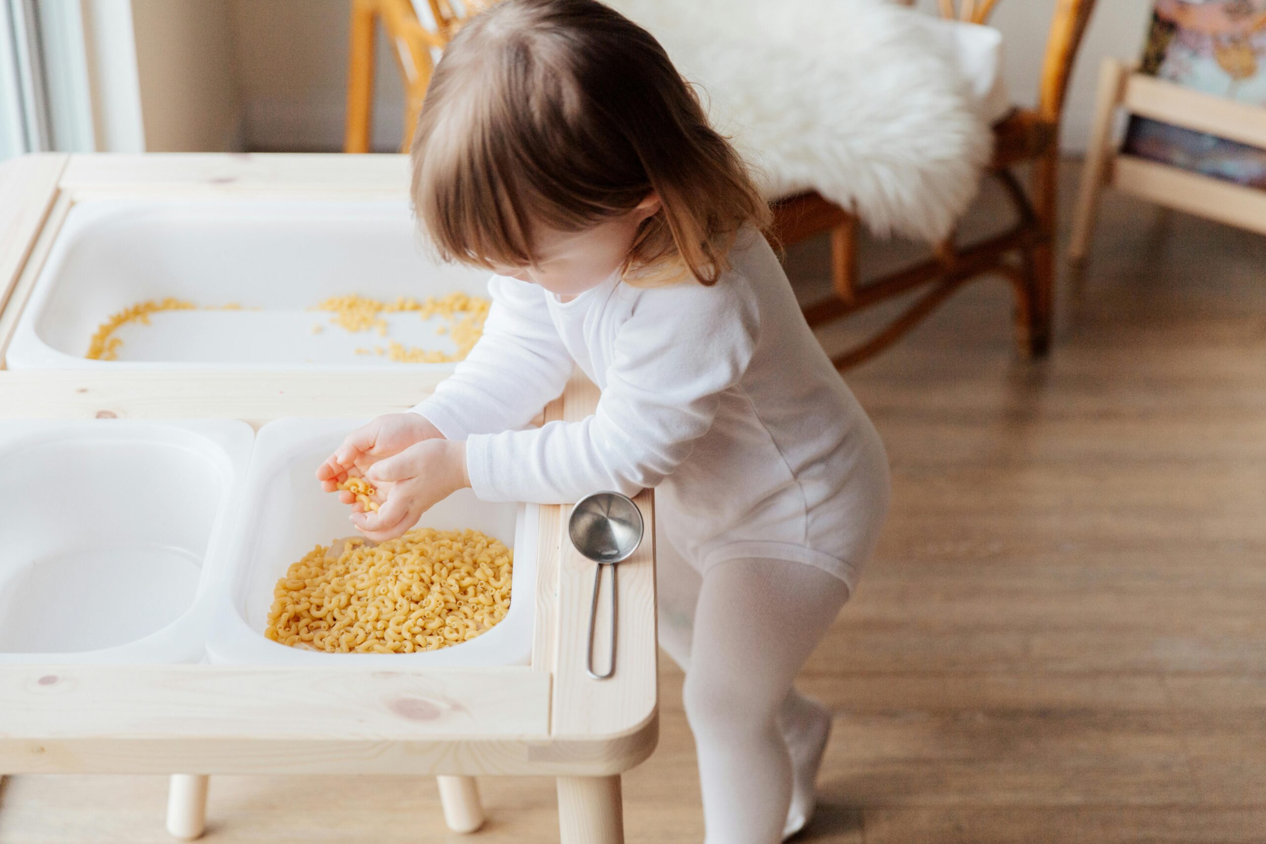 young two year old girl playing with uncooked pasta in a bin to promote her early childhood development & meet her play milestones in early childhood