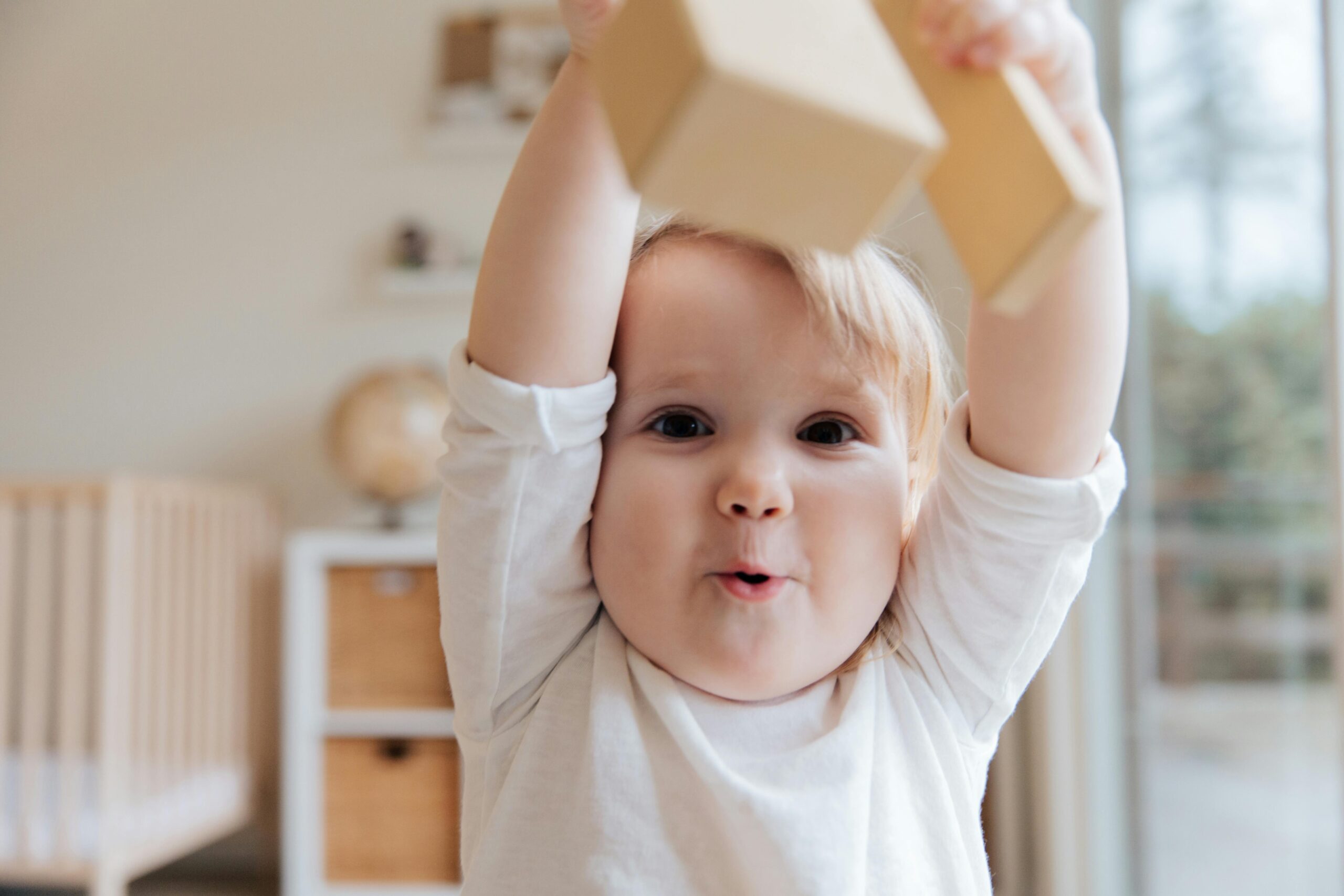 one year old baby looking directly at the camera while playfully lifting a wooden block while making noises to meet their play milestones in early childhood